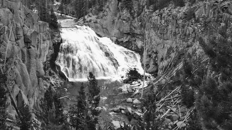 Waterfall tumbling over rocky ledges into a shallow pool, with stone walls and pine trees framing the narrow canyon in Yellowstone National Park.