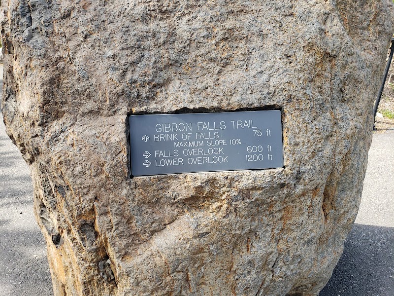 Large rock with a metal plaque labeled Gibbon Falls Trail and overlook directions in Yellowstone National Park.