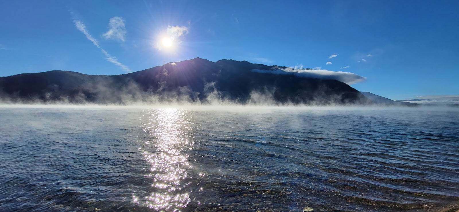 Fish Creek Picnic Area at Glacier National Park, sunlit lake with mist rising from the water and mountain silhouette in the distance.