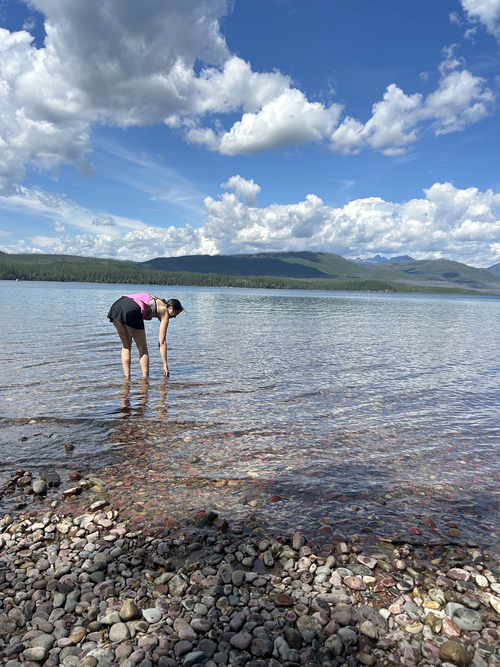 Person wading at Fish Creek Picnic Area along Glacier National Park’s shoreline with pebbled beach and blue sky.