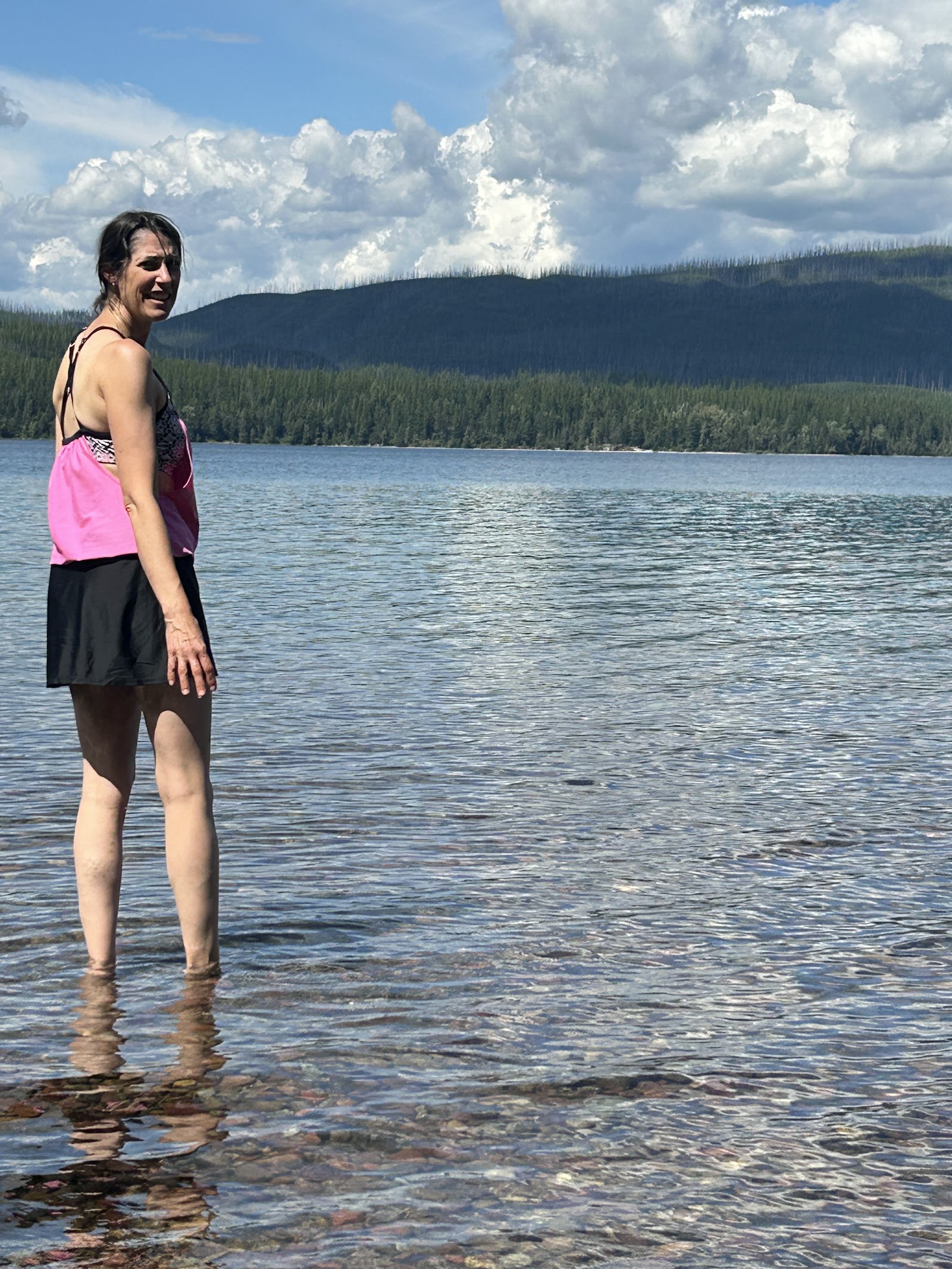 Visitor stands in shallow Fish Creek at the Fish Creek Picnic Area in Glacier National Park, with forested hills beyond.