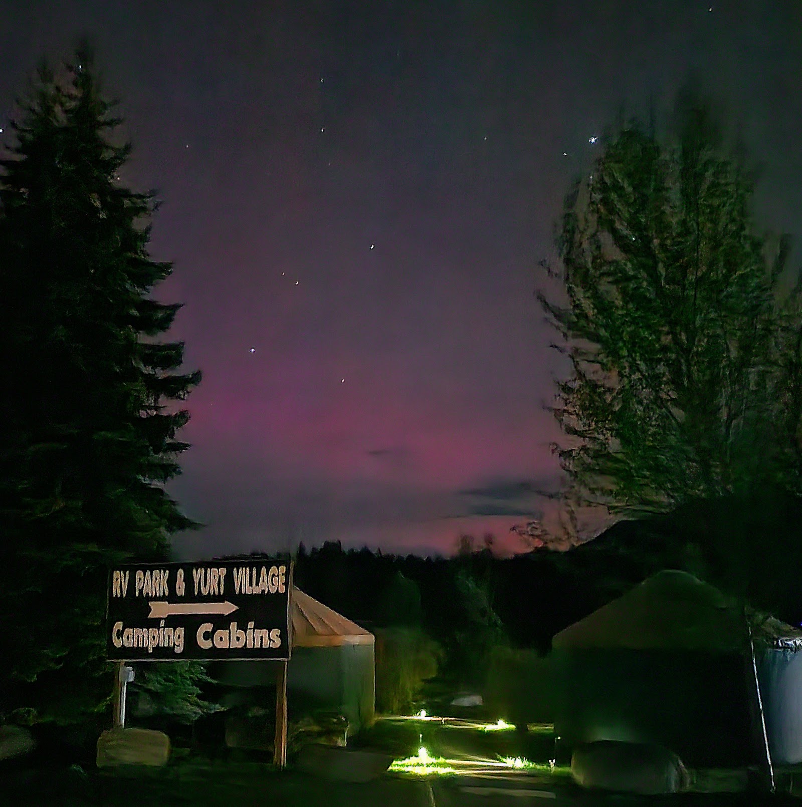 Night campground scene with yurts and tents among tall pines, glowing sign, and a purple twilight sky in Glacier National Park.