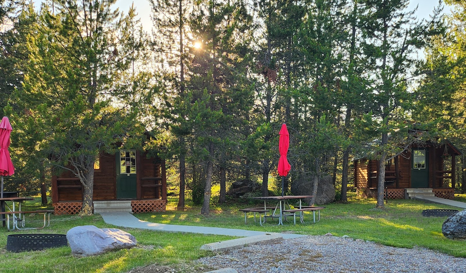 Wooden cabins with green doors amid pines, picnic tables, and red umbrellas at Glacier National Park campground.