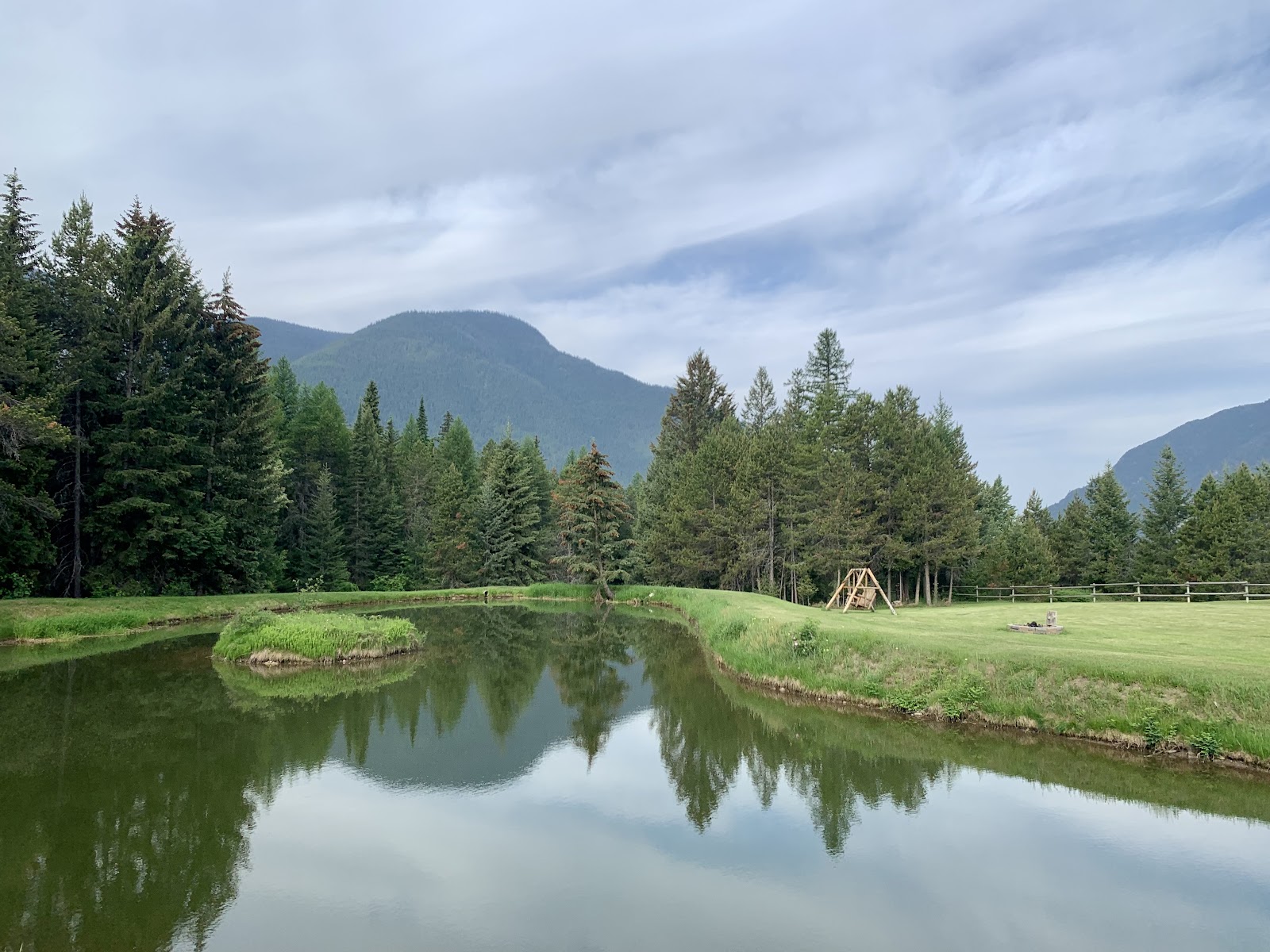 Calm pond in a grassy meadow reflects evergreen trees and distant mountains in Glacier National Park, with a wooden swing set and fence nearby.