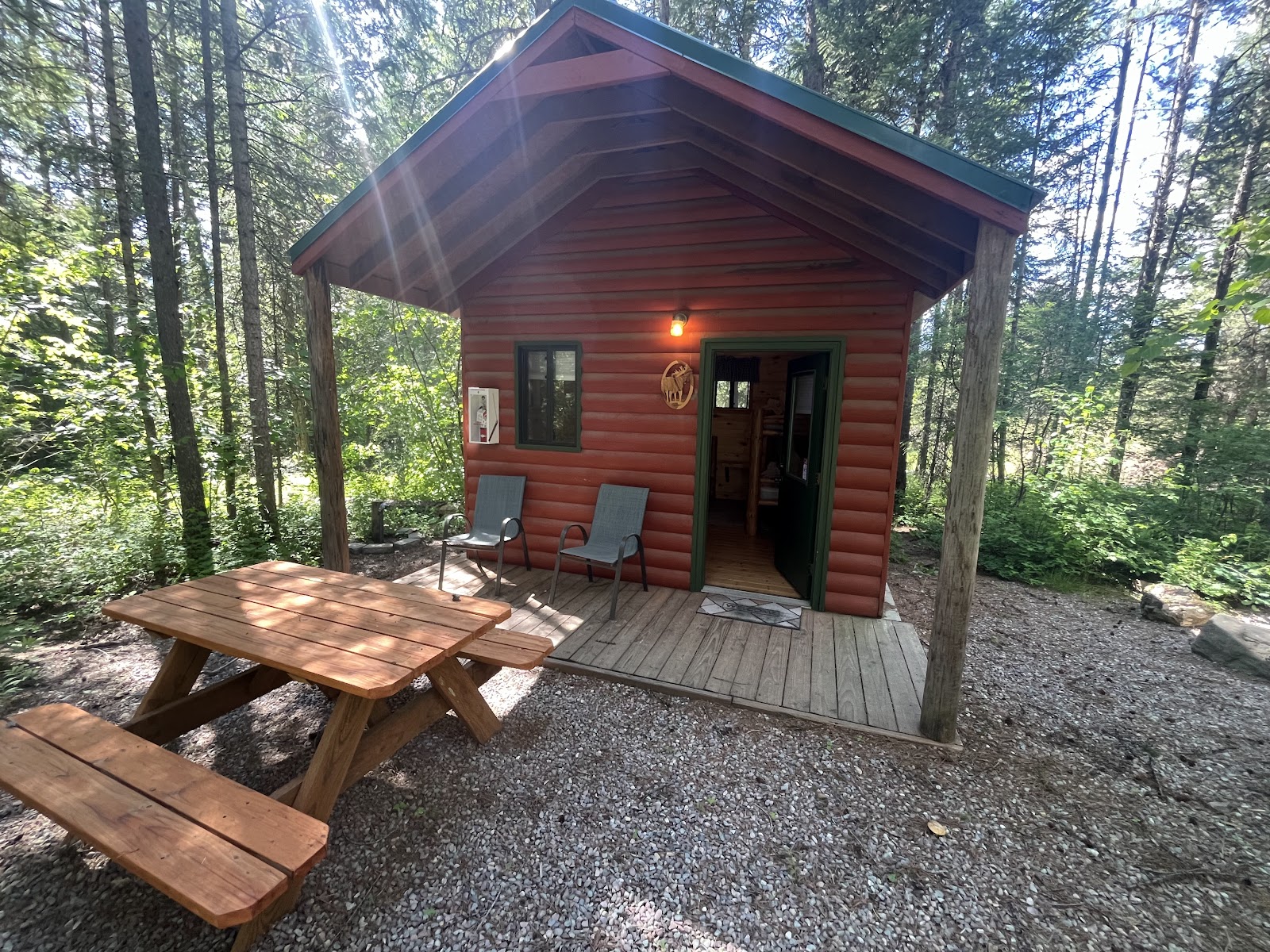 Red log cabin with green trim on a wooden deck in a pine forest, two chairs and a picnic table, Glacier National Park.