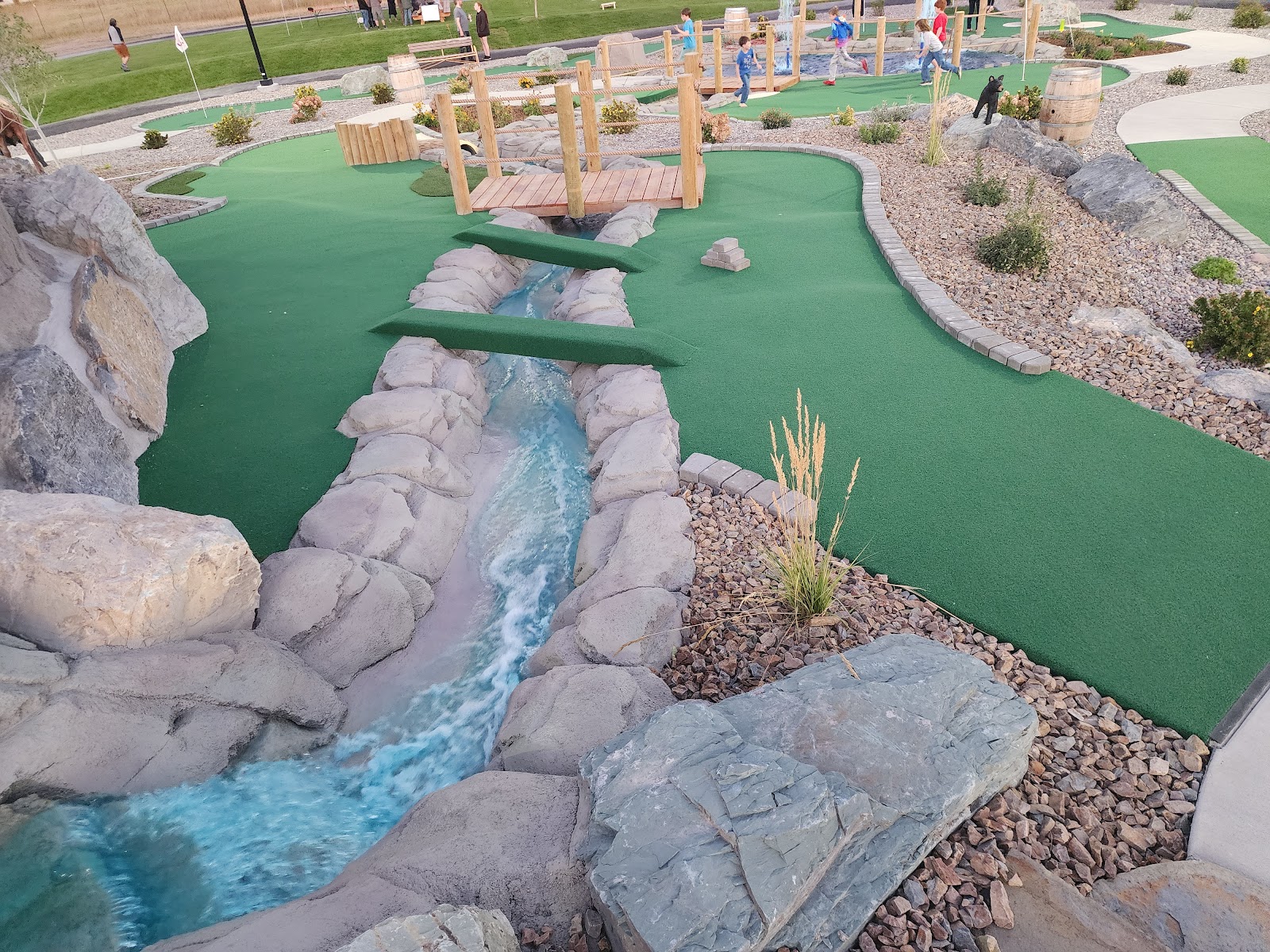 People play mini-golf on a synthetic green course with wooden bridge obstacles, a faux stream lined by rocks, and gravel landscaping in Glacier National Park.