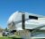 A large RV trailer sits at a Glacier National Park campground, with picnic tables nearby and a clear blue sky overhead.