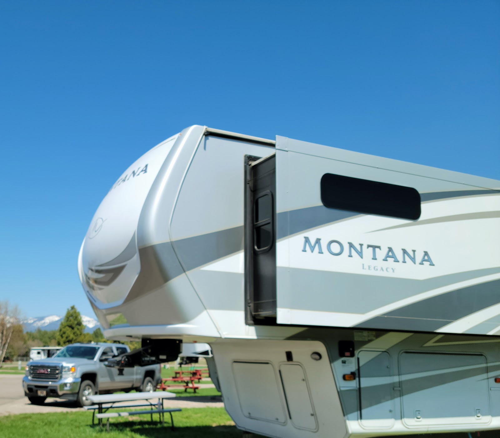 A large gray-and-white fifth-wheel RV sits at a campground in Glacier National Park, under a clear blue sky with green grass.
