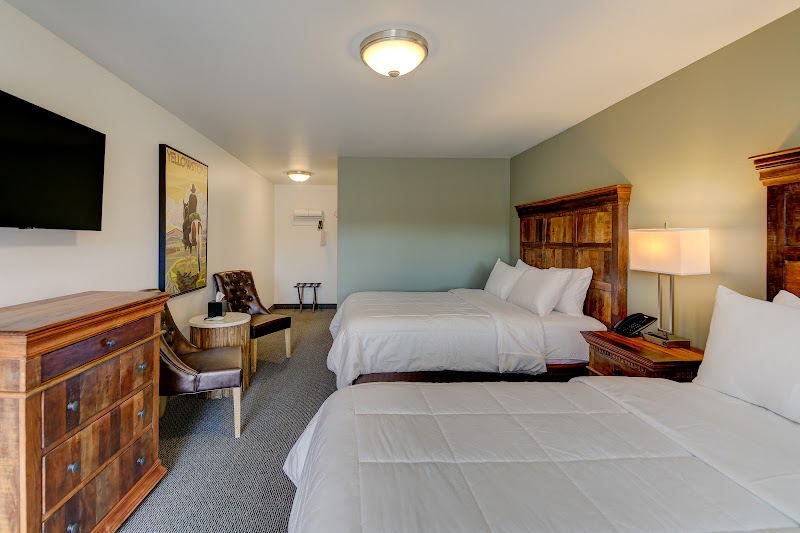 Two queen beds with wooden headboards, a dresser, wall-mounted TV, seating area, and a poster in a Yellowstone National Park hotel room.