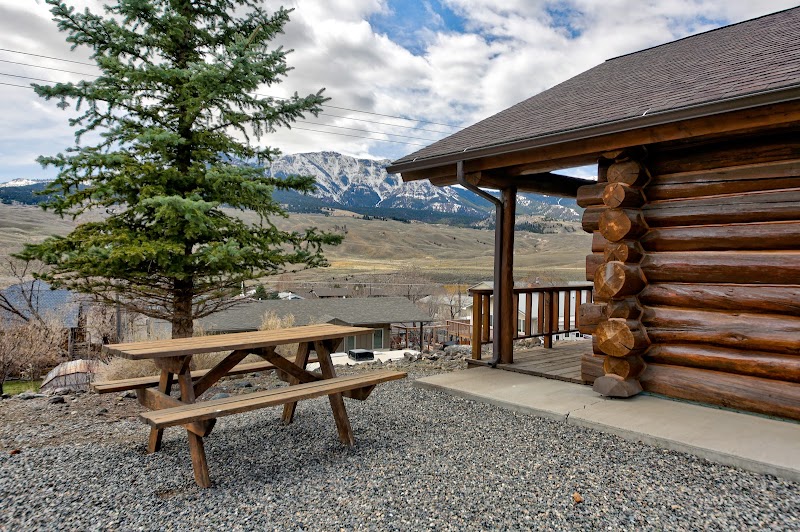 Log cabin with wooden porch and railing, gravel yard, picnic table, snow-capped mountains in Yellowstone National Park.
