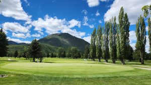 Green golf green with flags, surrounded by tall trees and a mountain in Glacier National Park under a bright blue sky with scattered white clouds.