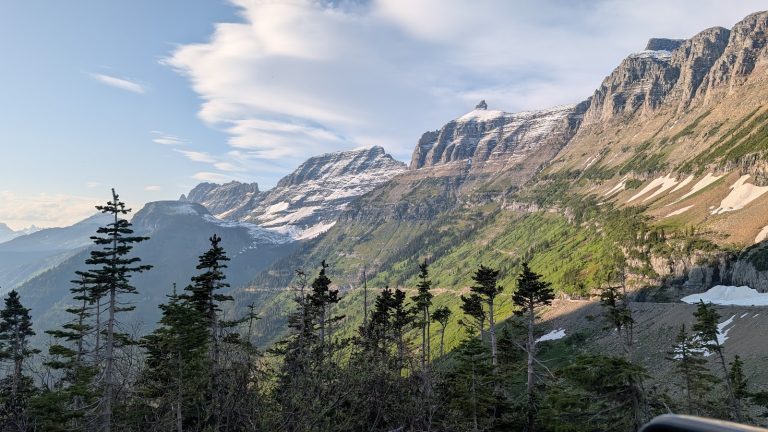 Snow-dusted jagged mountain peaks rise above green slopes and a pine forest in Glacier National Park.