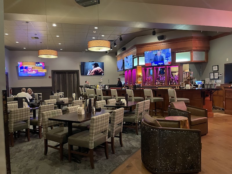 Cozy restaurant interior with a dark wood bar, purple backlit shelves, many tables with plaid chairs, and wall-mounted TVs in Yellowstone National Park.