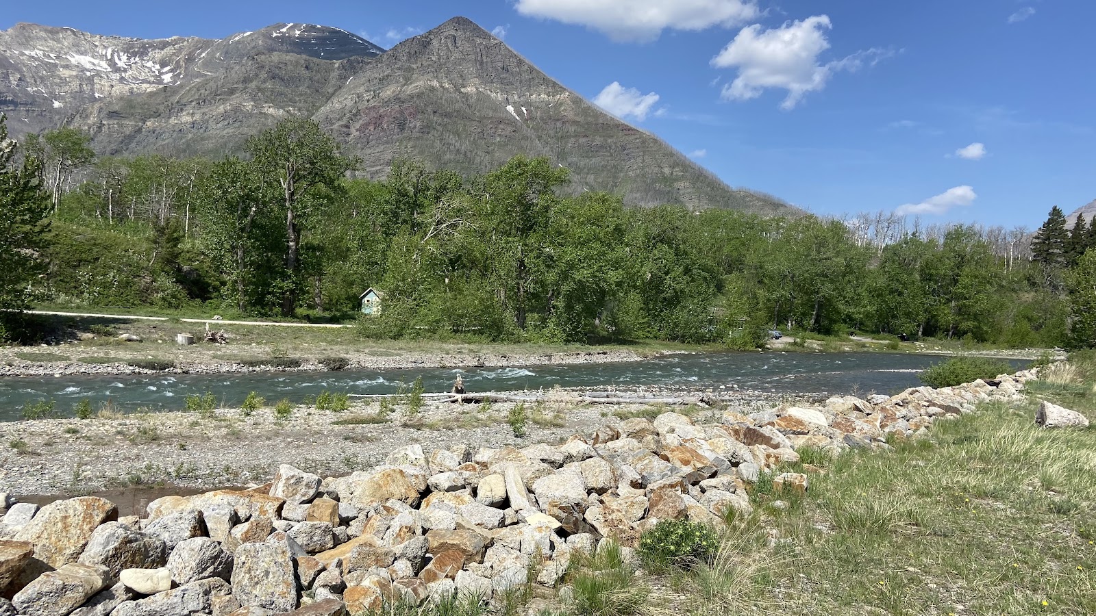 Pass Creek Day Use Area with a rocky riverbank, lush trees, and Glacier National Park mountains under a blue sky.