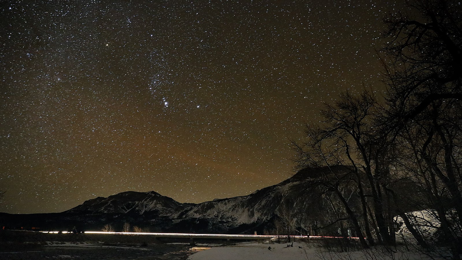 Night sky over Pass Creek Day Use Area in Glacier National Park, with snow-covered peaks and bare trees.