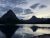 Jagged peaks rise over a calm glacier-fed lake with a rocky shore and tree line in Glacier National Park at dusk.