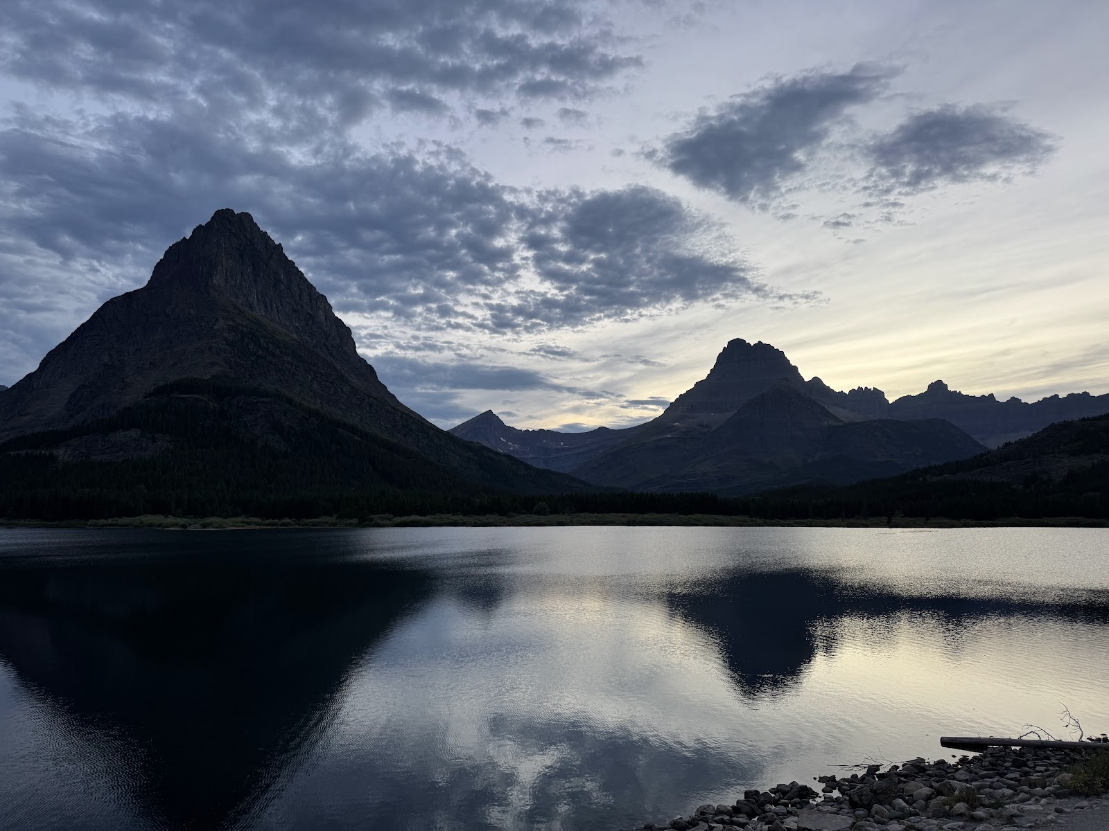 Jagged peaks rise over a calm glacier-fed lake with a rocky shore and tree line in Glacier National Park at dusk.