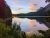 Sunset over a calm alpine lake at Glacier National Park, with evergreen shoreline, rocky left edge, and distant rugged mountains.