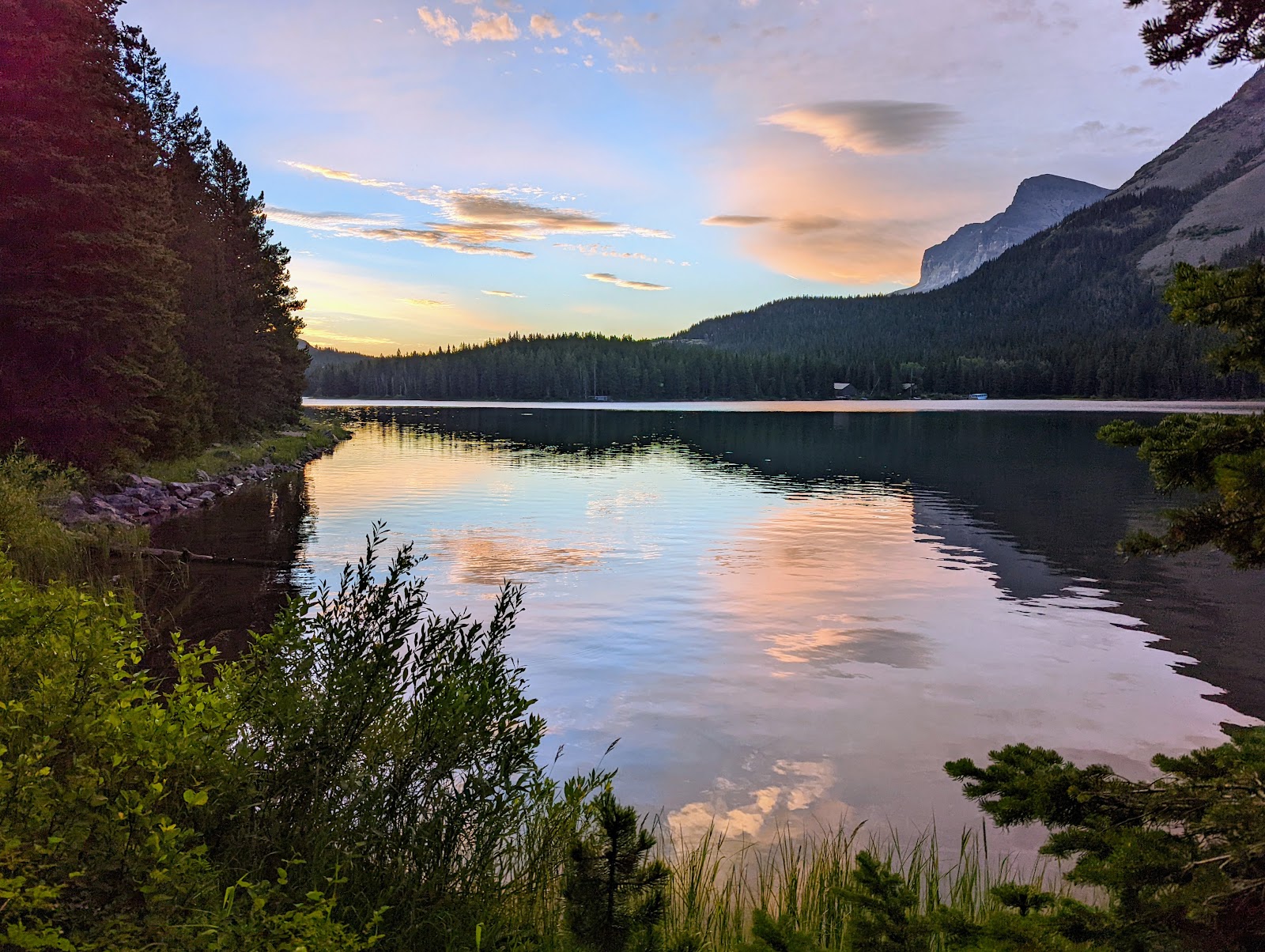 Sunset over a calm alpine lake at Glacier National Park, with evergreen shoreline, rocky left edge, and distant rugged mountains.
