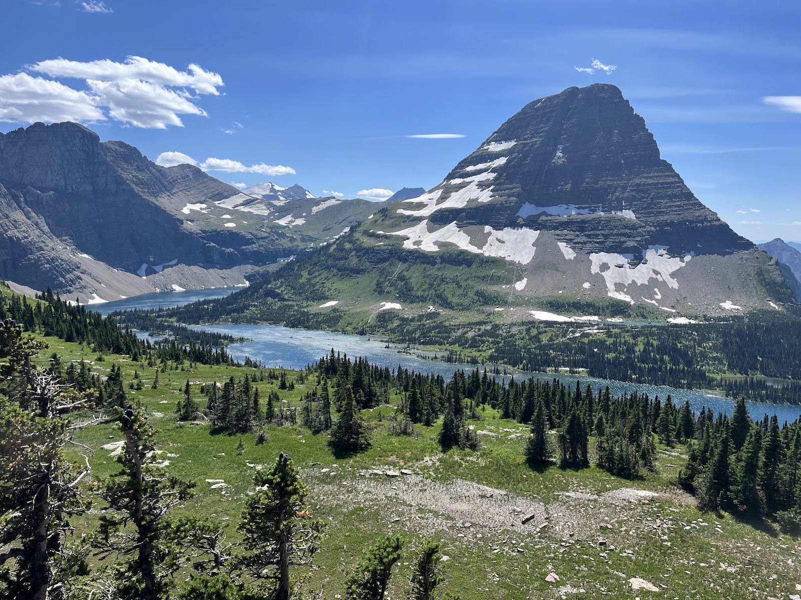 Snow-dusted peaks rise over a turquoise glacier-fed lake, meadows, and evergreen forests in Glacier National Park.