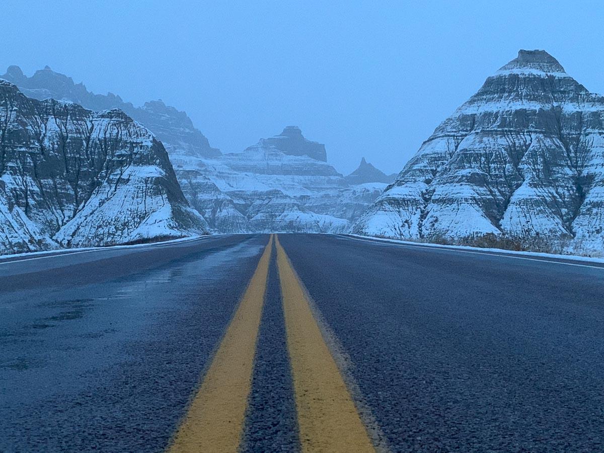 Snow-covered badlands rise on both sides as a two-lane road with yellow lines cuts through Badlands National Park.