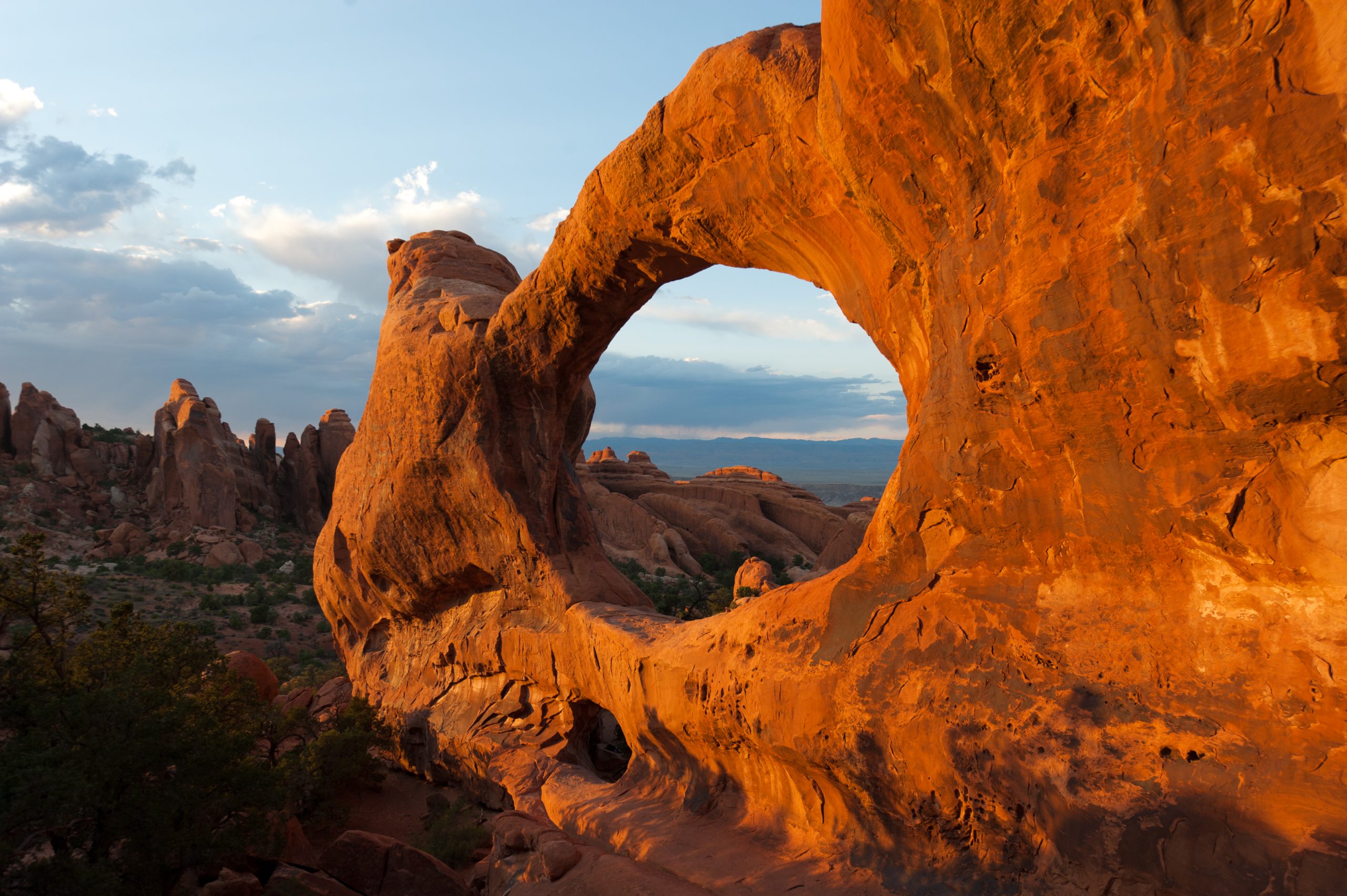 Sunset light bathes a large orange sandstone arch in Arches National Park, with jagged rock spires and blue sky beyond.
