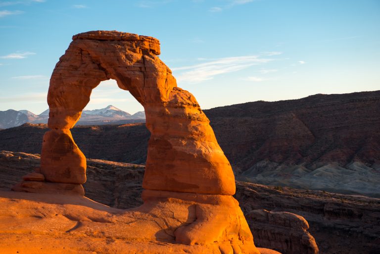 Delicate Arch in Arches National Park glows orange at sunset with distant desert ridges and mountains.