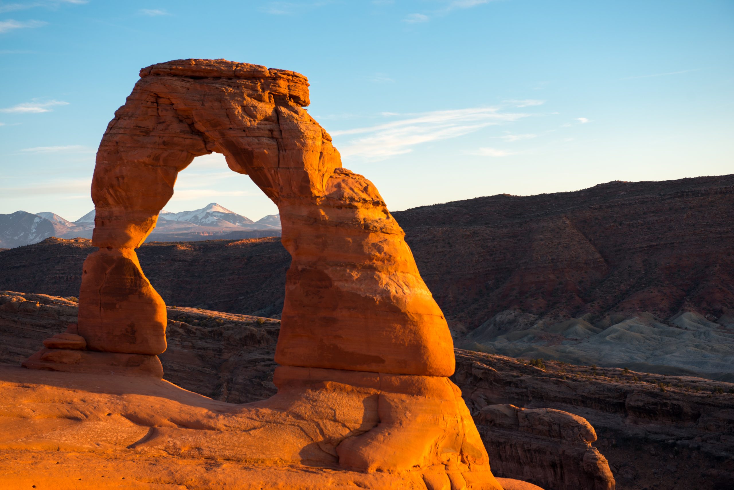 Delicate Arch stands in orange sandstone at Arches National Park, with a desert valley and distant snow-capped peaks.