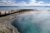 Wooden boardwalk extends over a steaming turquoise hot spring toward a calm lake against distant mountains in Grand Prismatic Spring, Yellowstone National Park.