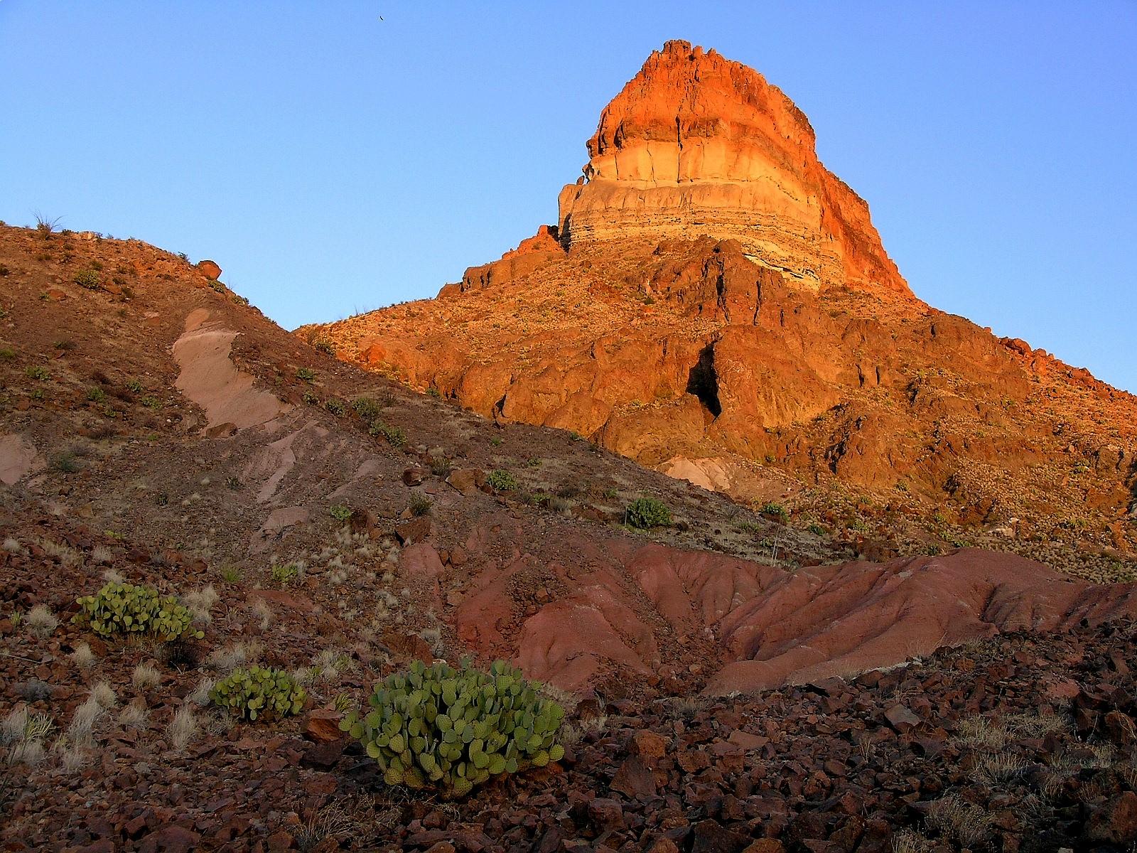 Red rock formation rises over rugged desert slopes in Big Bend National Park at golden hour.