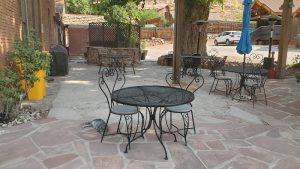 Outdoor stone patio with black wrought-iron tables and chairs, a blue umbrella, and a parked car near Arches National Park