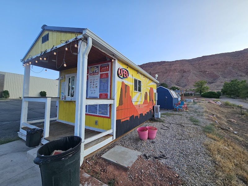 Bright yellow snack stand with a mural and open sign, menu posters, in Arches National Park, gravel lot, desert hills in the background.