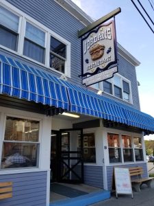 Blue two-story restaurant with a striped blue awning, large windows, open door, and a hanging sign at Acadia National Park.