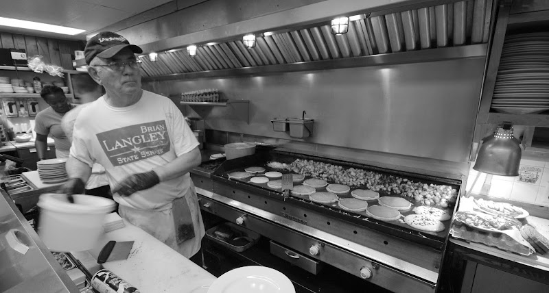 Busy diner kitchen in Acadia National Park with a cook flipping burgers on a griddle while plates rack and hood lights glow.