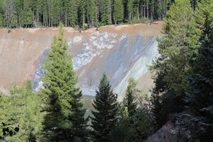 Essex area in Glacier National Park shows a colorful exposed hillside with pine trees at the rim and forest beyond.
