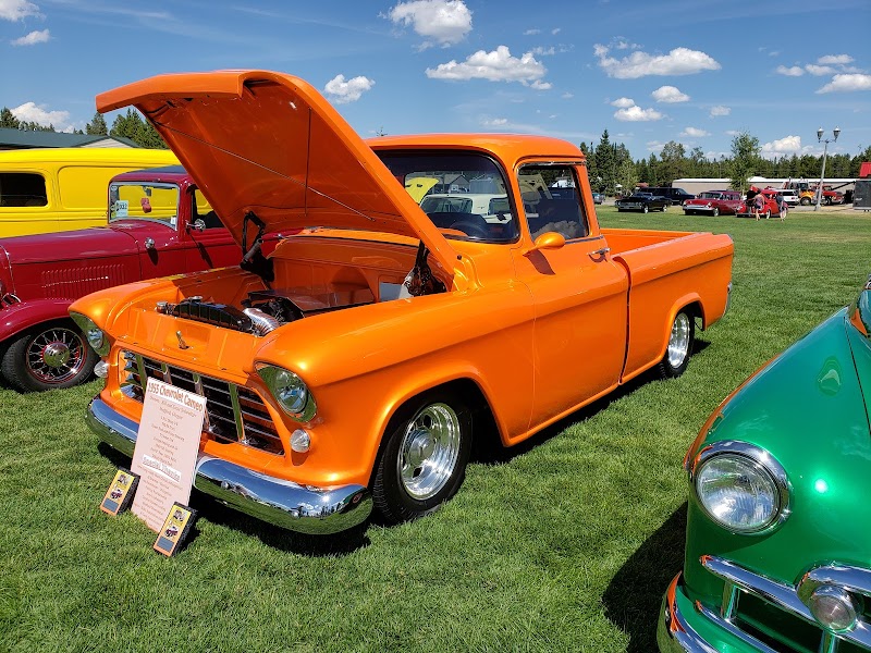 Bright orange vintage pickup with the hood open sits on a grassy field at Yellowstone National Park, beside colorful classic cars.