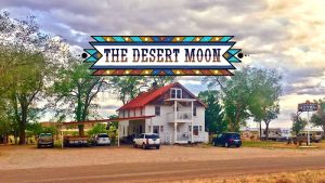 Two-story white house with a red roof sits at a dusty campground in Arches National Park, with several cars and an RV nearby.