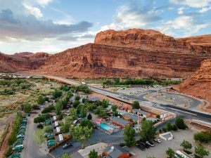 Campground rows of tents and cars with a winding road, set against red rock cliffs in Arches National Park.