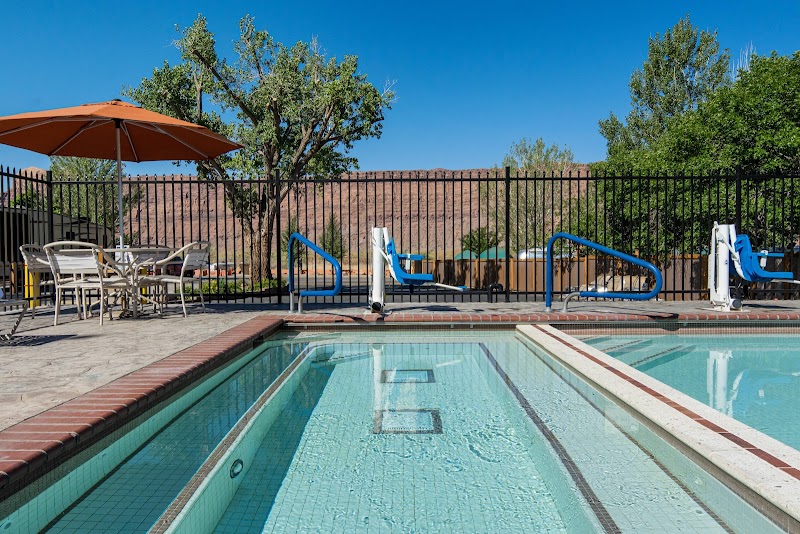 Poolside view with blue railing, orange umbrella, white chairs and tables, and desert hills near Arches National Park.