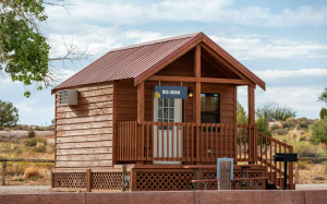 A wooden cabin with a front porch and railing sits in a desert campground at Arches National Park, under a blue sky.
