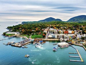A lively harbor in Acadia National Park with docks, boats, and a colorful town set against green hills and distant mountains.