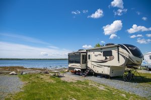 Bright day at Acadia National Park campground with a large beige RV by a rocky shoreline, bikes and a picnic table nearby.