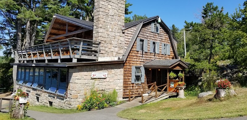 Stone and timber restaurant with a stone chimney, wraparound deck, gray shutters, and surrounding pines in Acadia National Park.