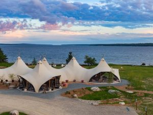 Glamping tents line a lakeside lawn with expansive ocean views inside Acadia National Park and pink sunset skies above.
