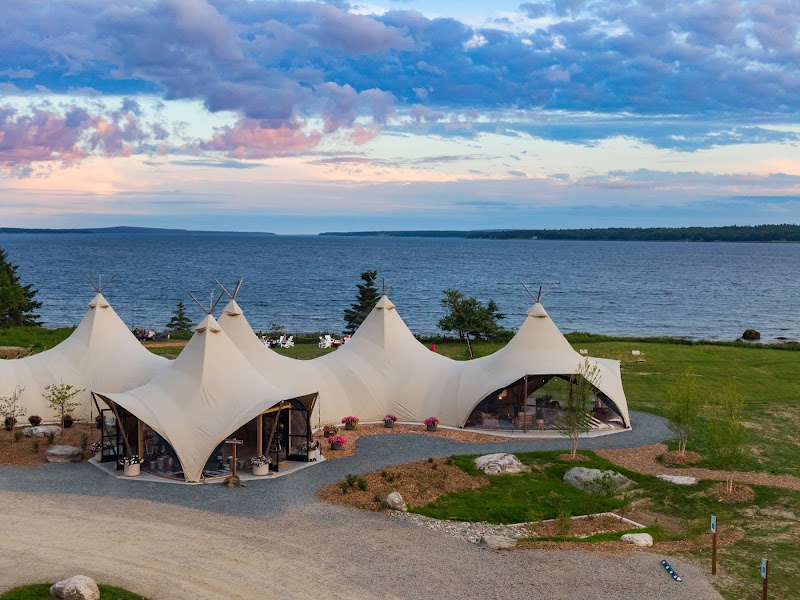 Glamping tents line a lakeside lawn with expansive ocean views inside Acadia National Park and pink sunset skies above.