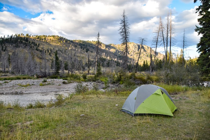Green and gray tent on a grassy riverbank with a rocky shore and pine-covered hills in Yellowstone National Park.