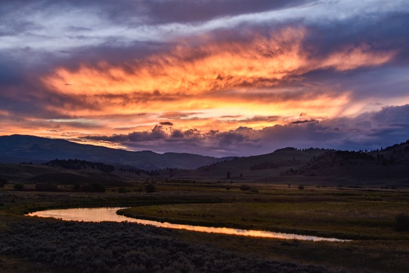 Fiery orange sunset over a winding creek through open grassland with distant hills in Yellowstone National Park.