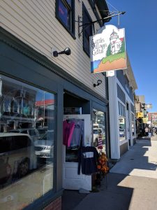 Sunlit storefront of a gift shop on Acadia National Park street, featuring a lighthouse sign and clothing display outside.