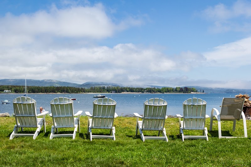 Six Adirondack chairs on a grassy shore overlook calm blue water with boats and forested hills in Acadia National Park.