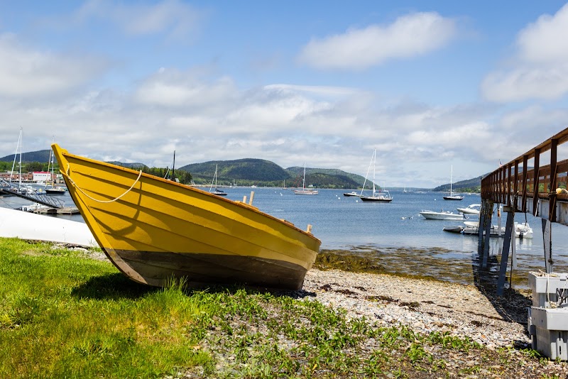 Yellow rowboat rests on grassy, pebbly shore beside a wooden dock, with calm harbor and boats at Acadia National Park.